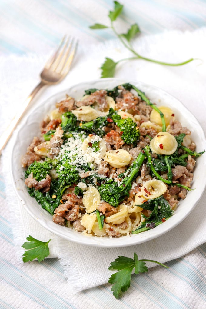 A white bowl filled with orecchiette pasta with sausage and broccoli rabe. A small bowl of grated parmesan in the background