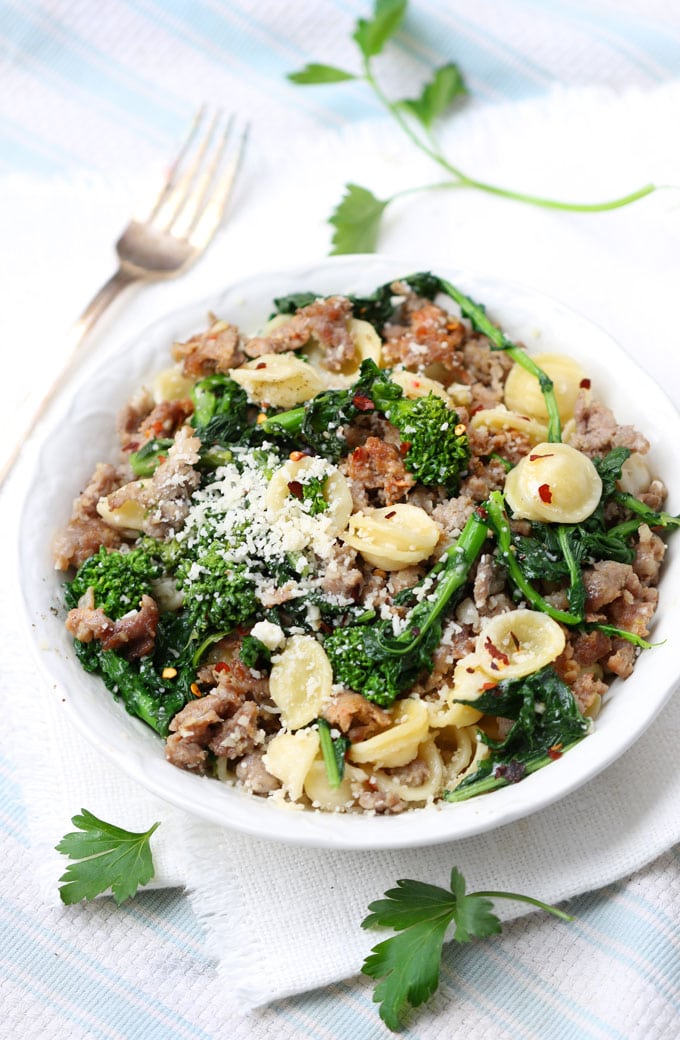 A white bowl seen from above, filled with orecchiette pasta tossed with a sauce of broccoli rabe and sausage. On the side is a fork a 3 sprigs of parsley.