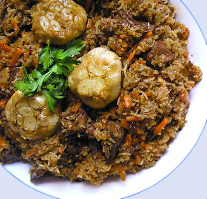 Plov: Lamb and rice and whole roasted bulbs of garlic in a bowl, garnished with parsley.