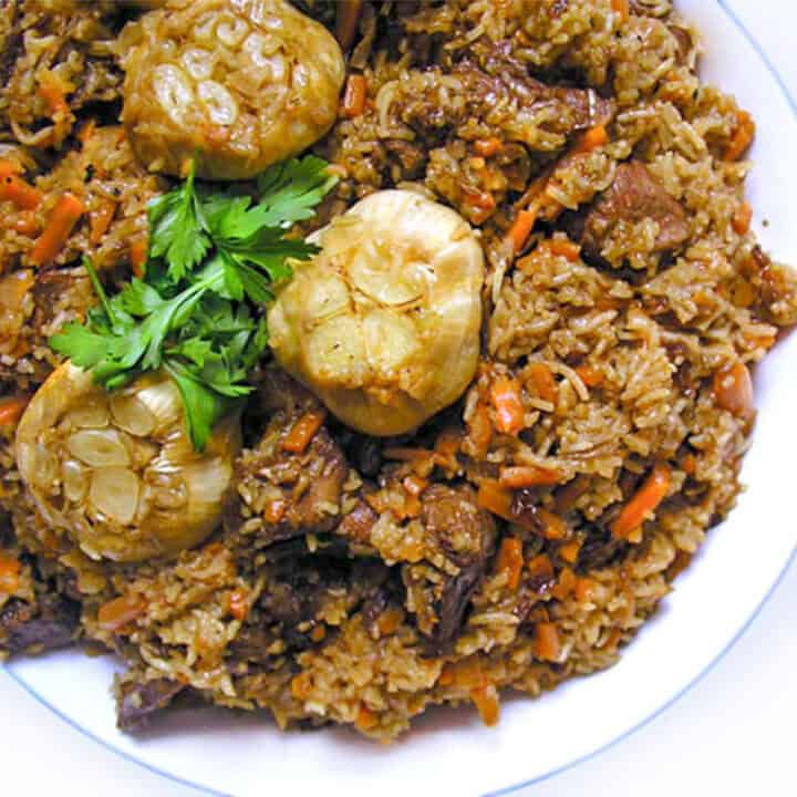 Uzbeki Plov on a plate - Rice pilaf with carrots and hunks of lamb in a bowl, with two roasted heads of garlic that have the tops sliced off.