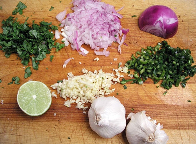 close up of a cutting board with piles of chopped ingredients: cilantro, red onion, garlic, and green chilies