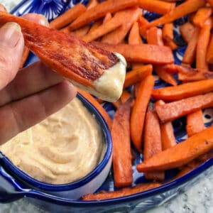 Sweet potato fries in a shallow serving bowl with chipotle aioli dip and one fry that has just been dipped, in someones fingers.