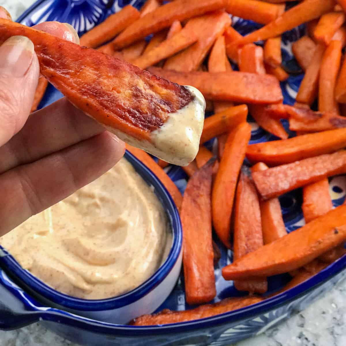 Sweet potato fries in a shallow serving bowl with chipotle aioli dip and one fry that has just been dipped, in someones fingers.