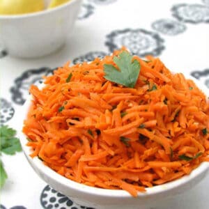 Shredded carrot salad in a bowl topped with a sprig of parsley. A bowl of lemons on the background.
