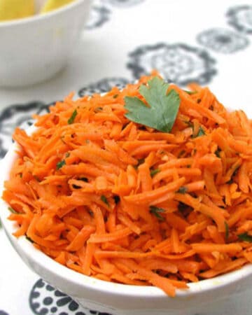 Shredded carrot salad in a bowl topped with a sprig of parsley. A bowl of lemons on the background.