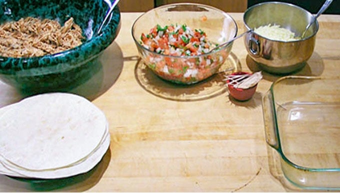 Bowls of ingredients for making BBQ chicken Burritos, on a kitchen counter: a large green bowl filled with shredded chicken, a glass bowl filled with homemade salsa, a metal bowl with cheese, a stack of flour tortillas and some toothpicks