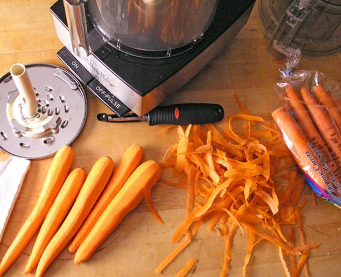 peeled carrots and carrot peelings on a counter next to a food processor and the shredding blade