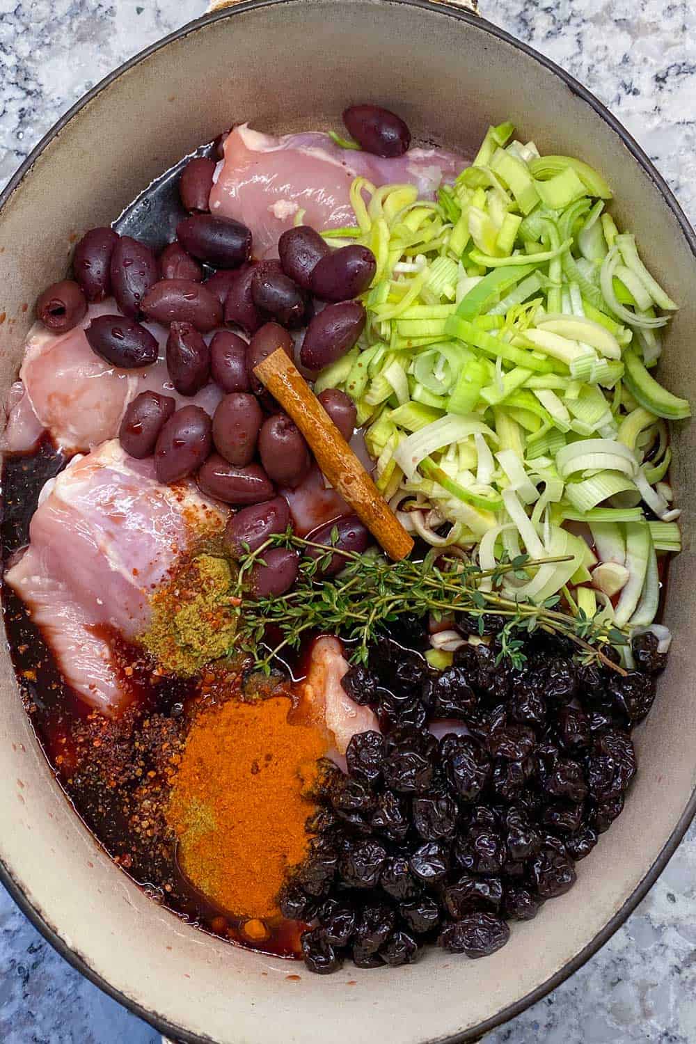 An oval White Dutch oven seen from above, with all the ingredients for slow cooker chicken thighs with dried cherries, leeks, kalamata olives and spices