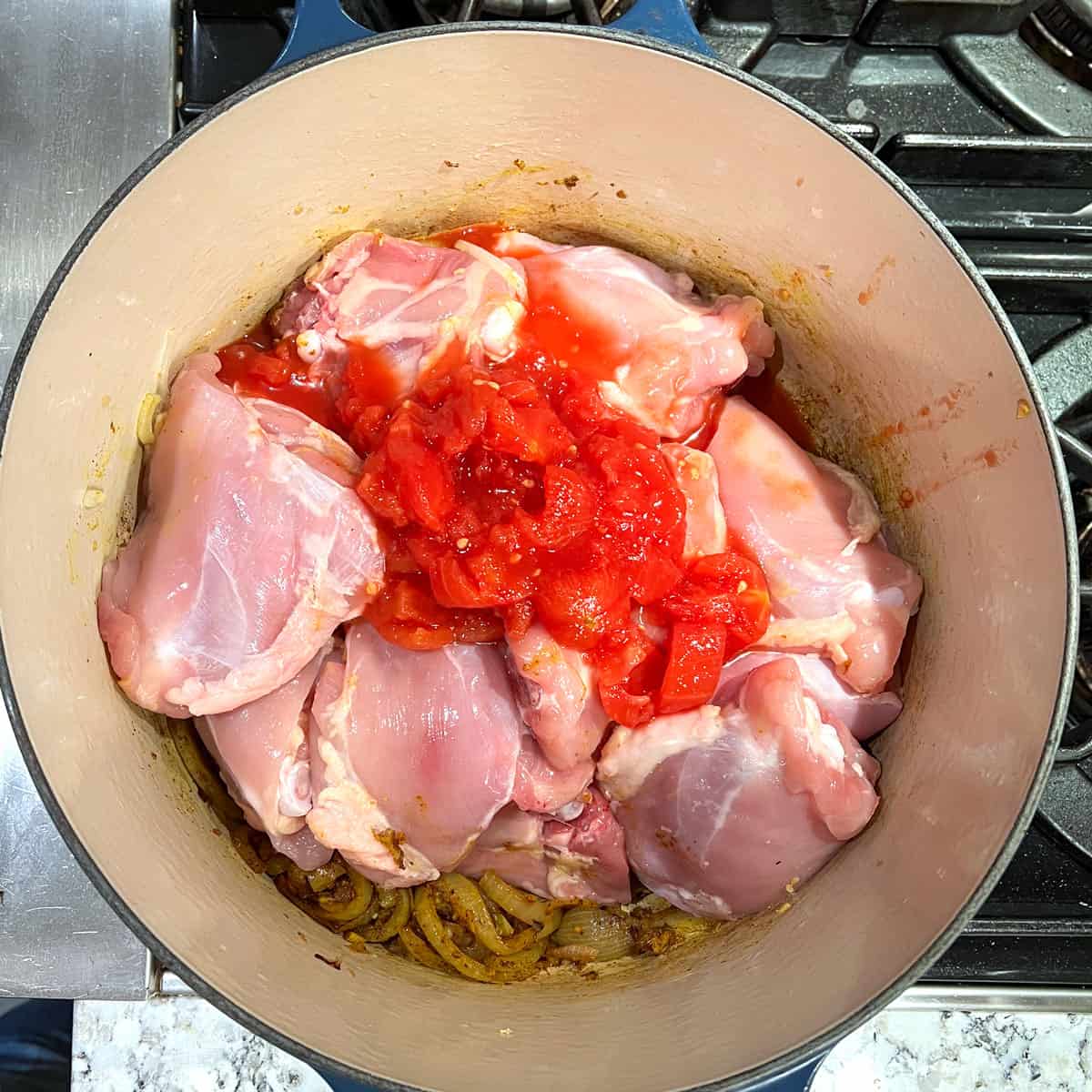 bone-in chicken thighs in a dutch oven on a bed of sauteed onions with canned chopped tomatoes on top.
