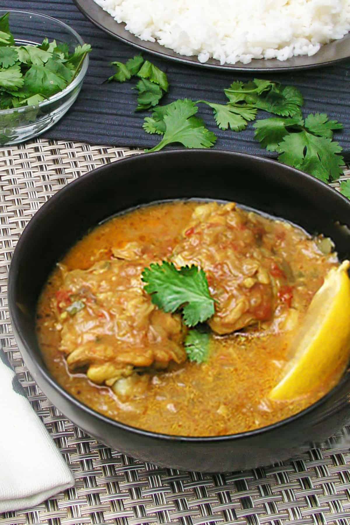 Bowl of coconut curry chicken with two chicken thighs in lots of coconut curry sauce, a lemon wedge in the bowl and a sprig of cilantro in the center.