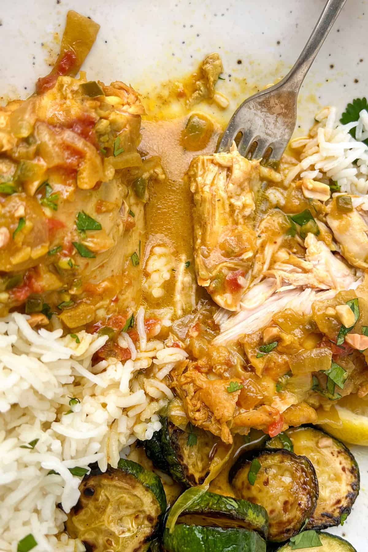 close up of coconut chicken thighs in a bowl with rice and sautéed zucchini, a fork pull at the tender chicken meat.