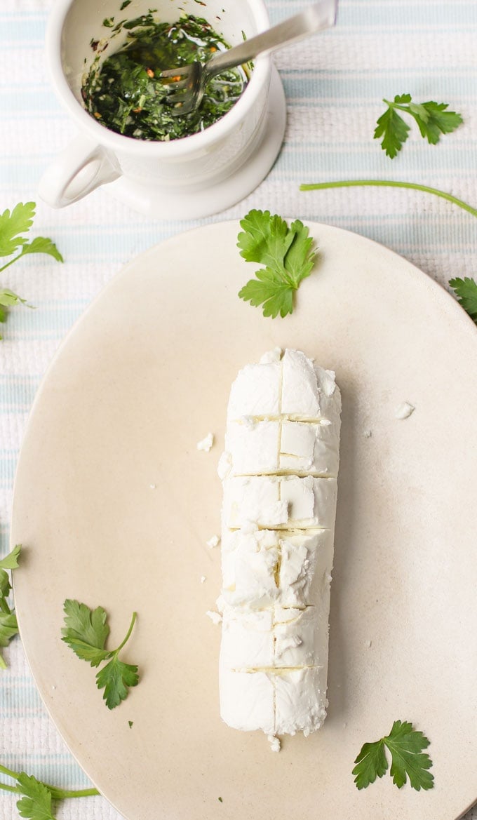 A log of goat cheese on a plate next to a cup of parsley dressing with parsley leaves sprinkled around