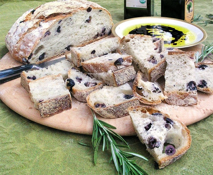 a round wooden serving board topped with a loaf of homemade no-knead olive bread that is partially sliced, with a small shallow bowl of olive oil and balsamic vinegar for dipping