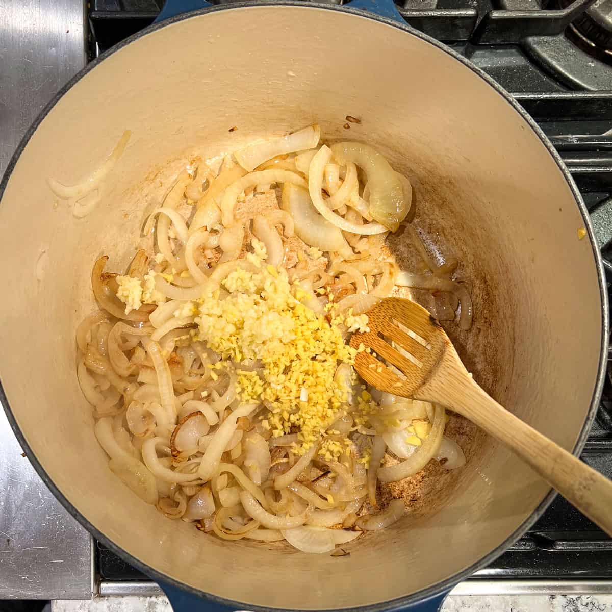 Wooden spoon stirring chopped garlic and ginger into sauteed onions in the bottom of a dutch oven.