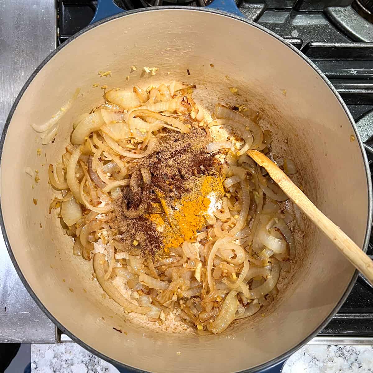 Spices added to a Dutch oven full of sauteed onions, a wooden spoon stirring.