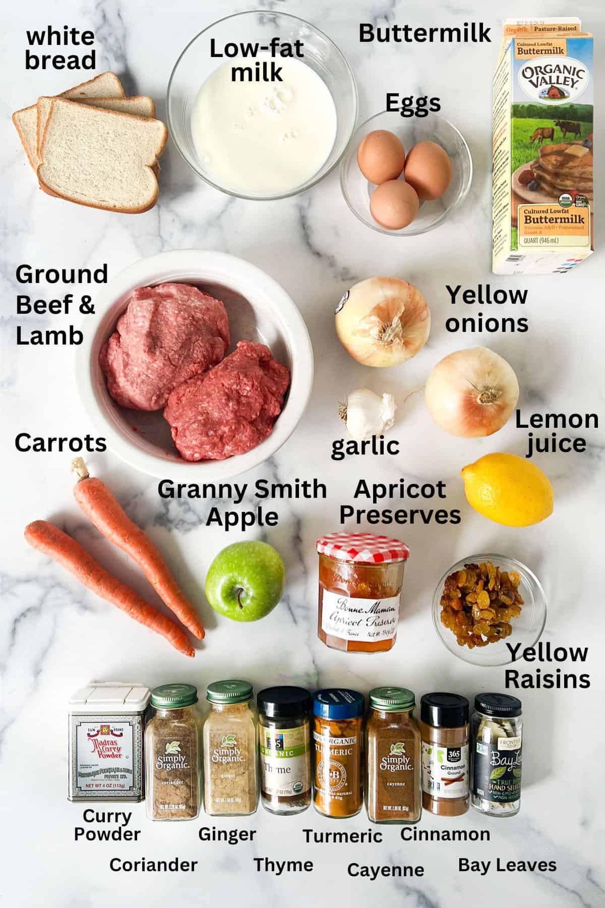 All the ingredients for Bobotie set out on a marble countertop: bread, milk, eggs, beef and lamb, lemon, onions, carrots, green apple, apricot preserves, garlic, yellow raisins, jars of spices.