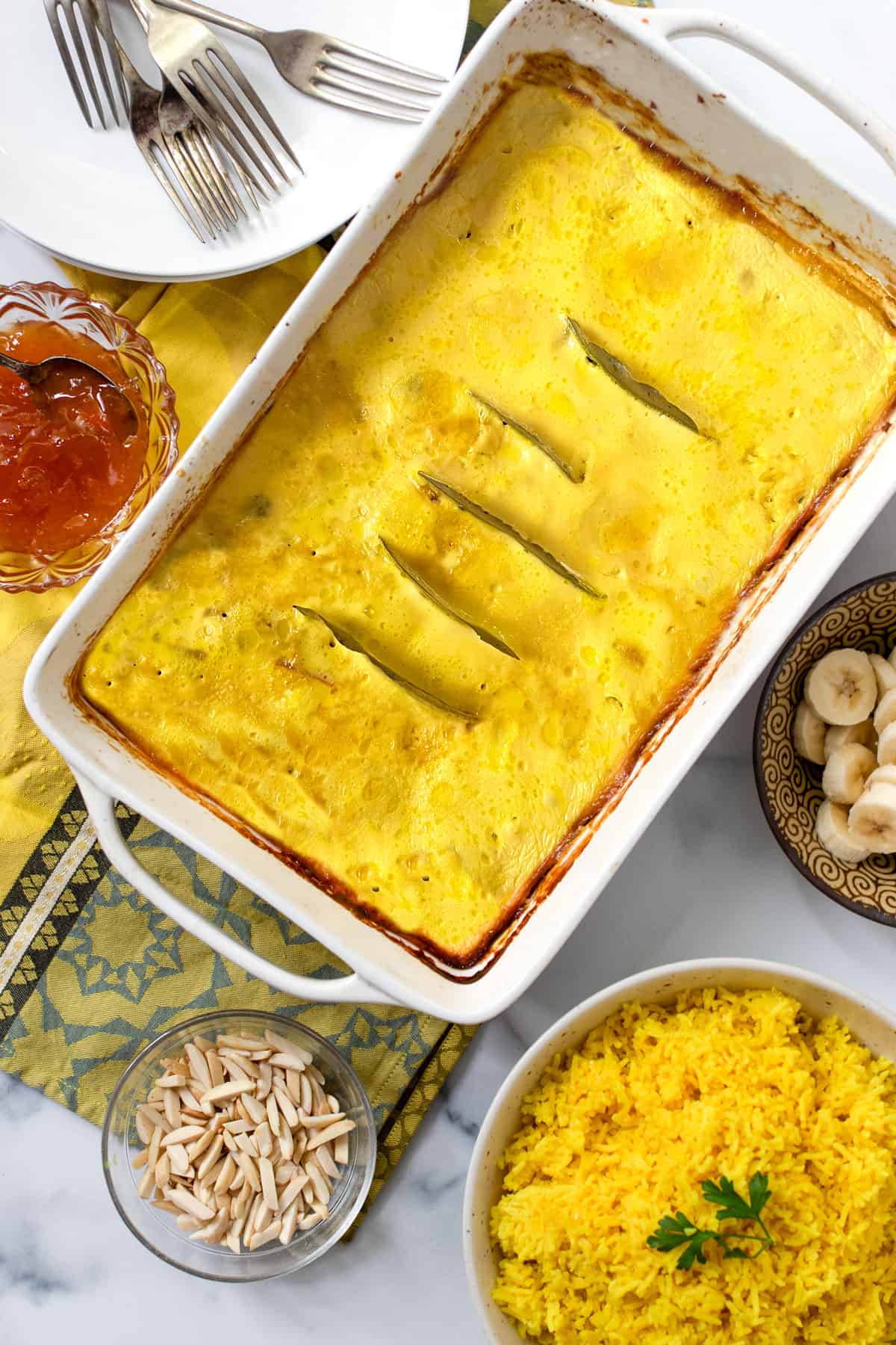 Rectangular casserole filled with custard topped Bobotie, bowls on the side of yellow rice, toasted slivered almonds, apricot preserves and sliced bananas.