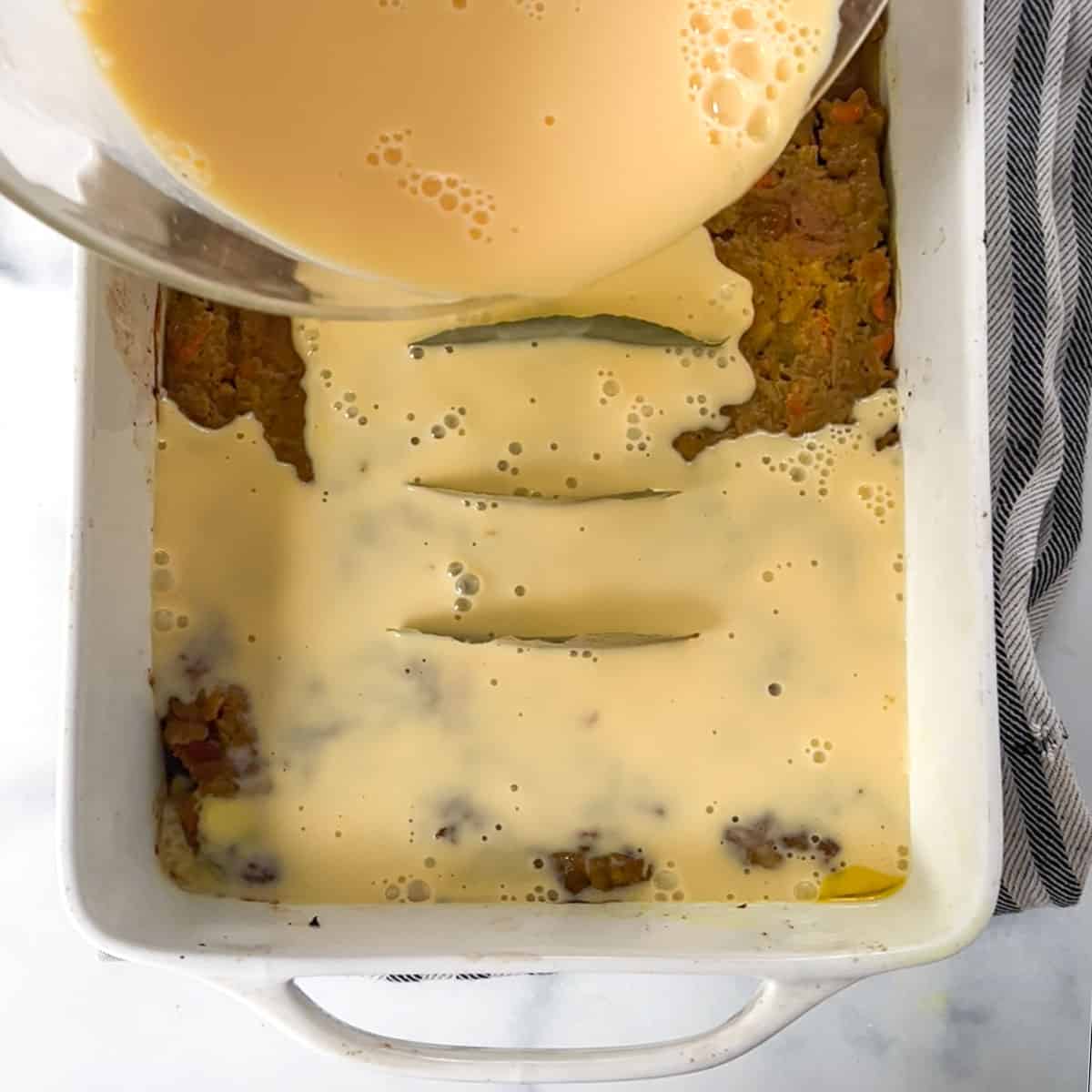 custard mixture being poured from a glass mixing bowl onto meat in a casserole.