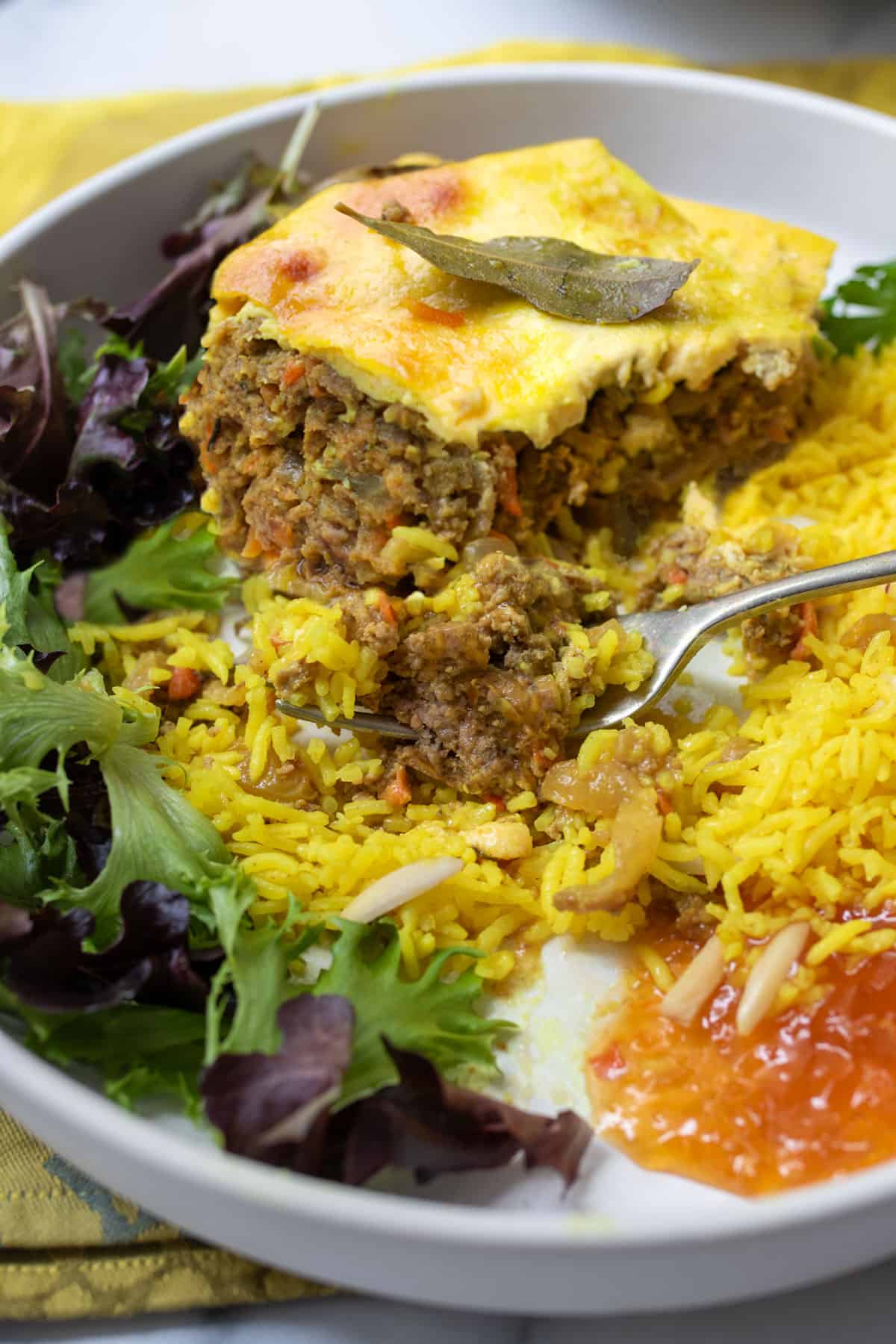 square slice of bobotie on a plate with yellow rice, salad and a blob of apricot preserves