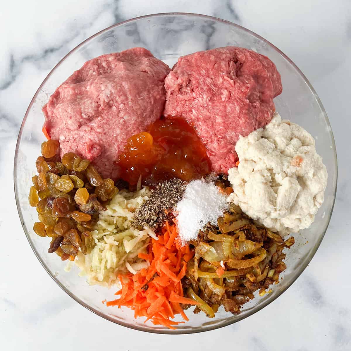 Glass bowl with ingredients for bobotie in separated piles: ground lamb, ground beef, yellow raisins, grated apples and carrots, sauteed onions with spices, milk-soaked bread, salt, pepper, apricot preserves.