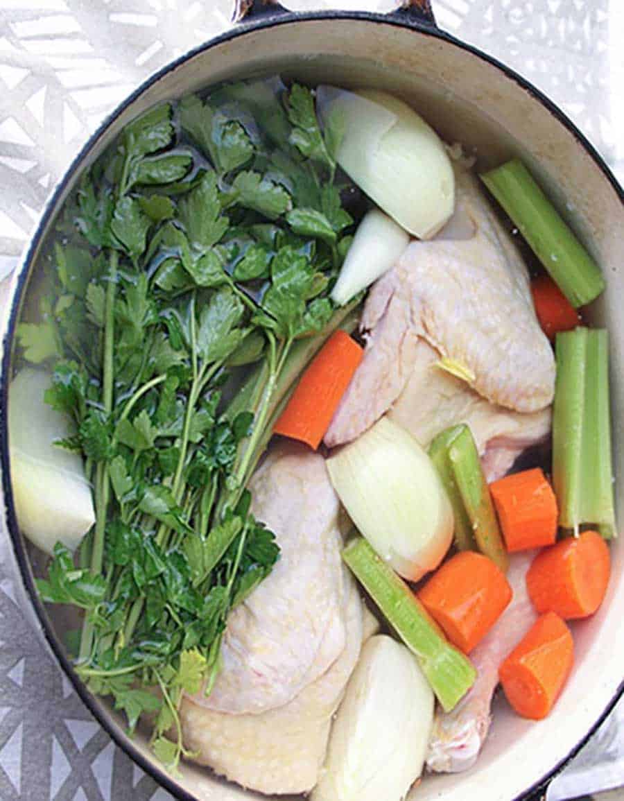 white oval Dutch oven filled with ingredients to make chicken soup from scratch: raw chicken, onions, carrots, celery and parsley