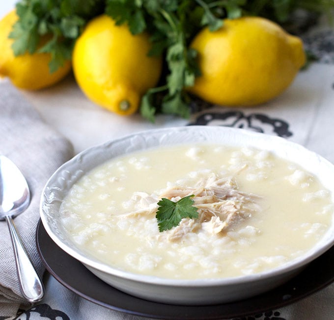 White bowl filled with Greek lemon chicken soup, shredded chicken in the middle and a parsley leaf, 3 lemons and a bunch of parsley in the background