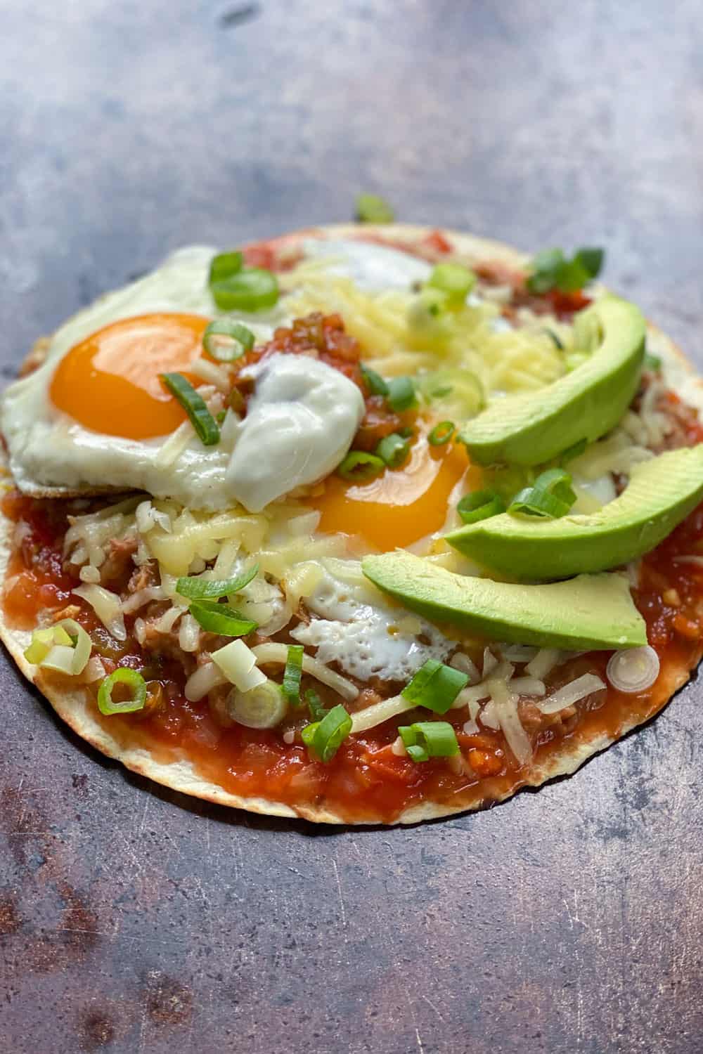 huevos rancheros: a flour tortilla on a sheet pan topped with salsa, refried beans, two sunny side up eggs, grated cheese, sliced avocados and chopped scallions.