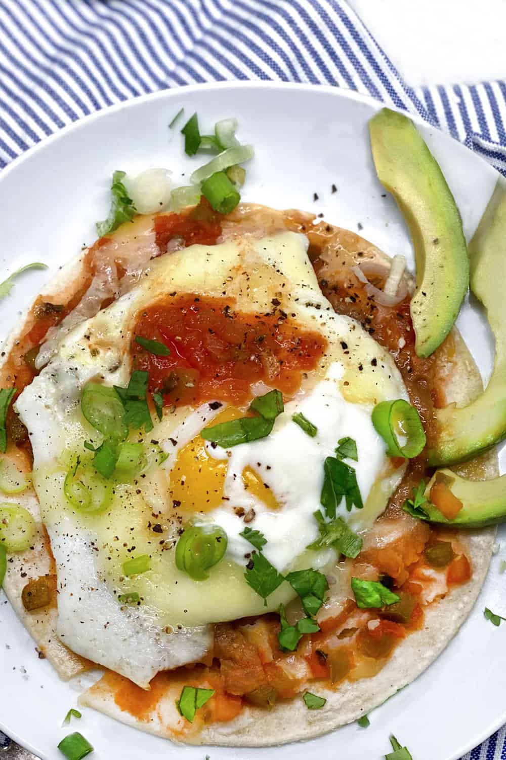 a white plate topped with huevos rancheros: a corn tortilla topped with refried beans, a gried egg, salsa, melted cheese, chopped scallions, a dollop of sour cream, and three slices of avocado.