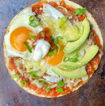 huevos rancheros on a copper colored baking sheet: a flour tortilla topped with salsa, refried beans, two fried eggs, shredded cheese and three slices of avocado