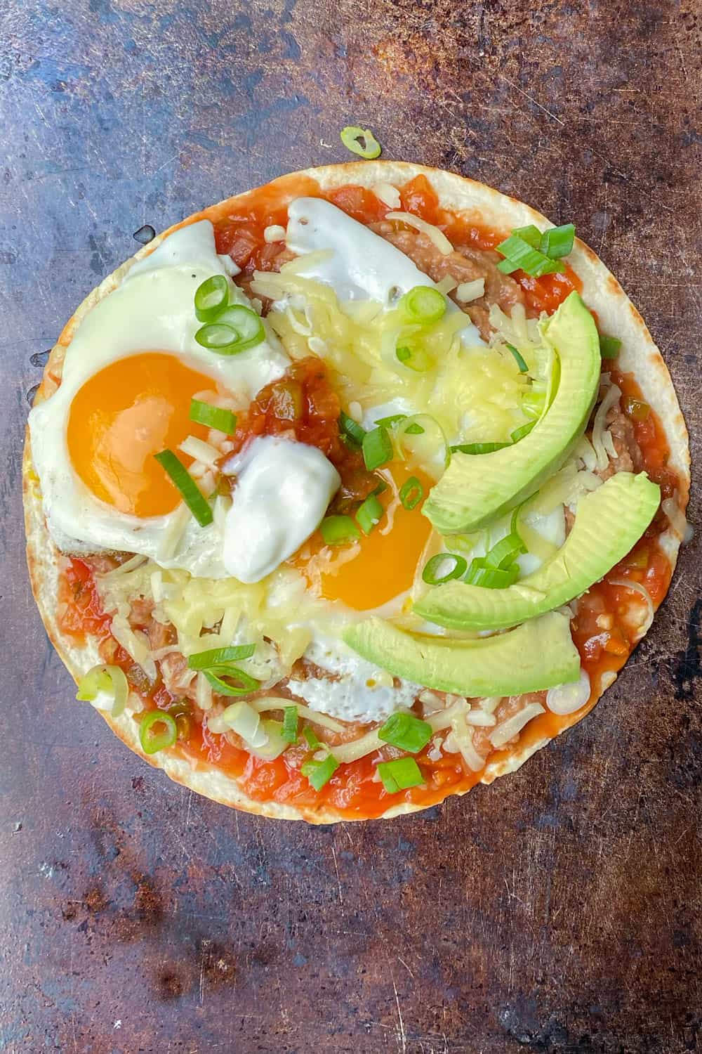 huevos rancheros on a copper colored baking sheet: a flour tortilla topped with salsa, refried beans, two fried eggs, shredded cheese and three slices of avocado