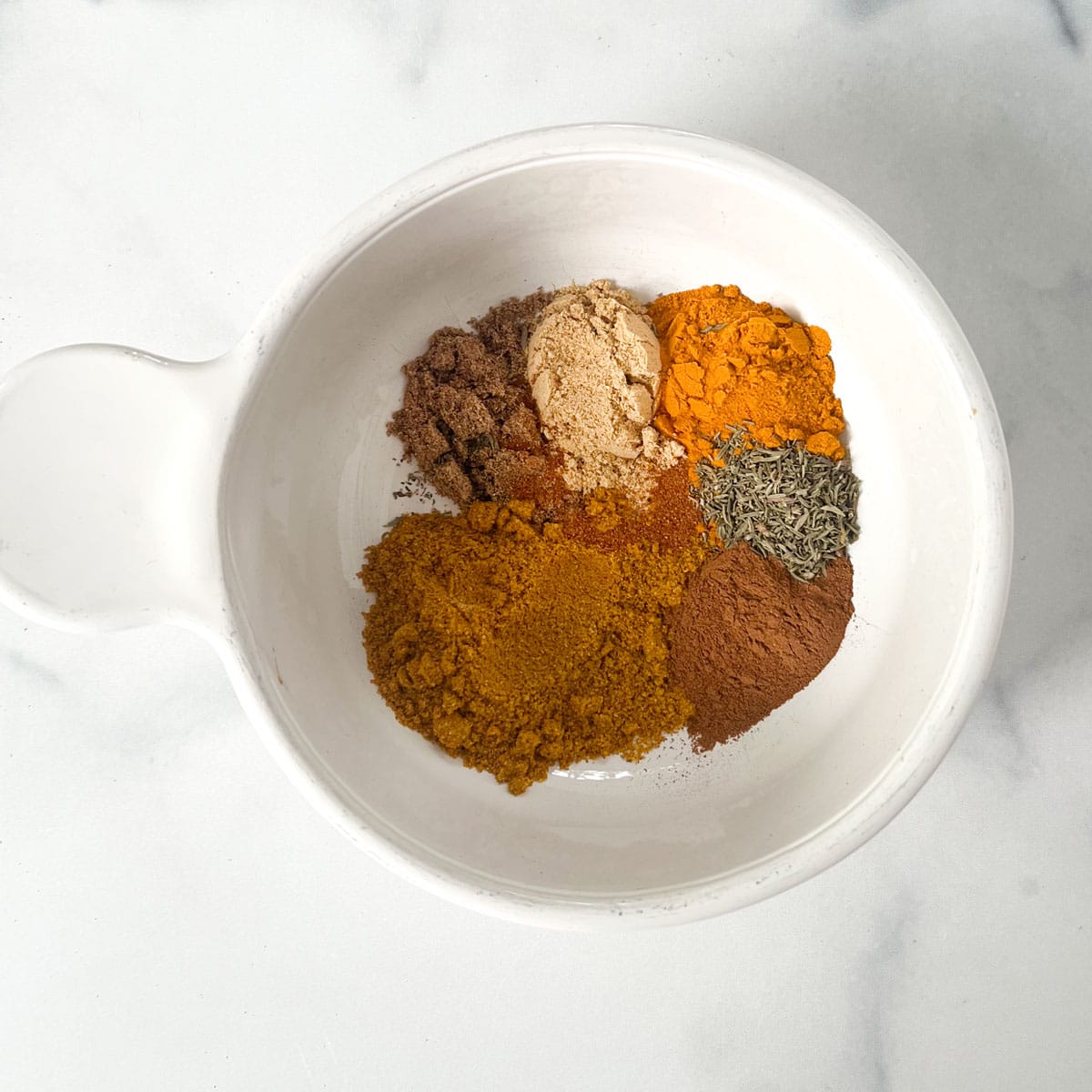 Small piles of ground spices in a bowl: curry powder, coriander, ginger, thyme, turmeric, cinnamon and cayenne.