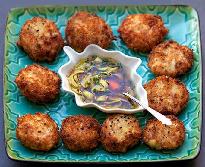10 crisp golden brown crab cakes on a green square platter with a small leaf-shaped bowl of sweet and sour dipping sauce in the center