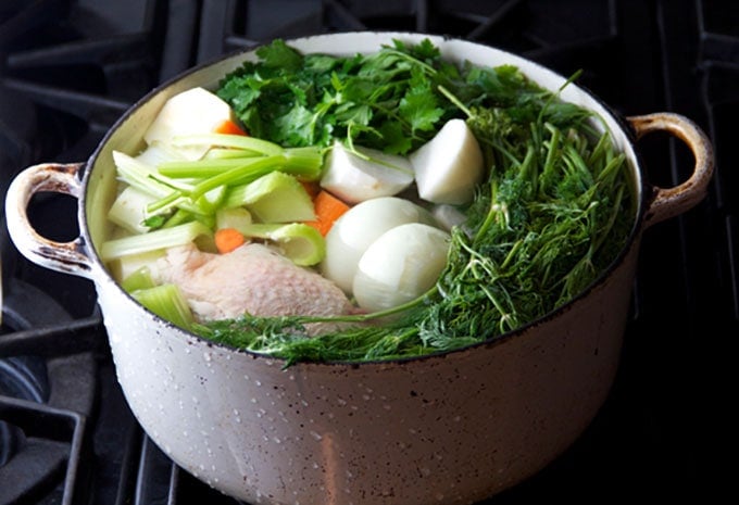 a white dutch oven filled to the brim with chicken, veggies and herbs, ready to make chicken soup