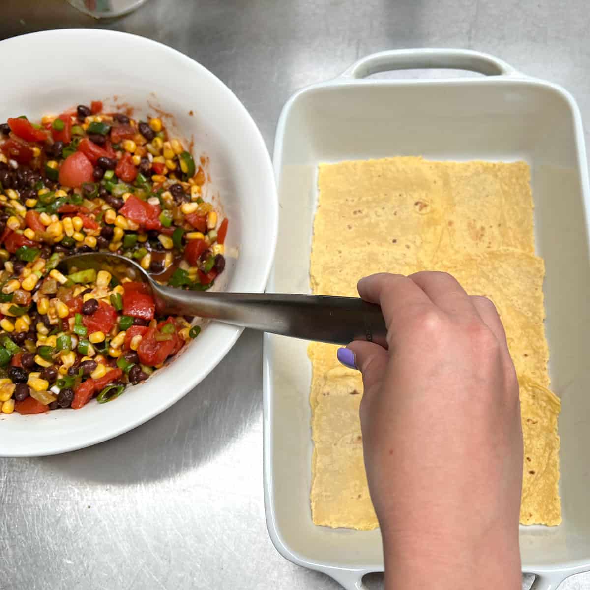 Baking pan fully lined with corn tortillas and a hand about to spoon in the first layer of bean, tomato and corn filling from a big bowl.