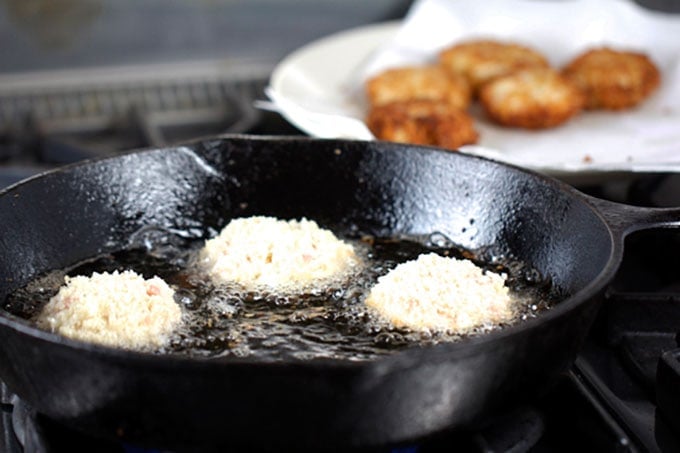 3 crab cakes sautéing in a cast iron skillet with 5 golden brown cooked crab cakes on a paper towel lined plate in the background