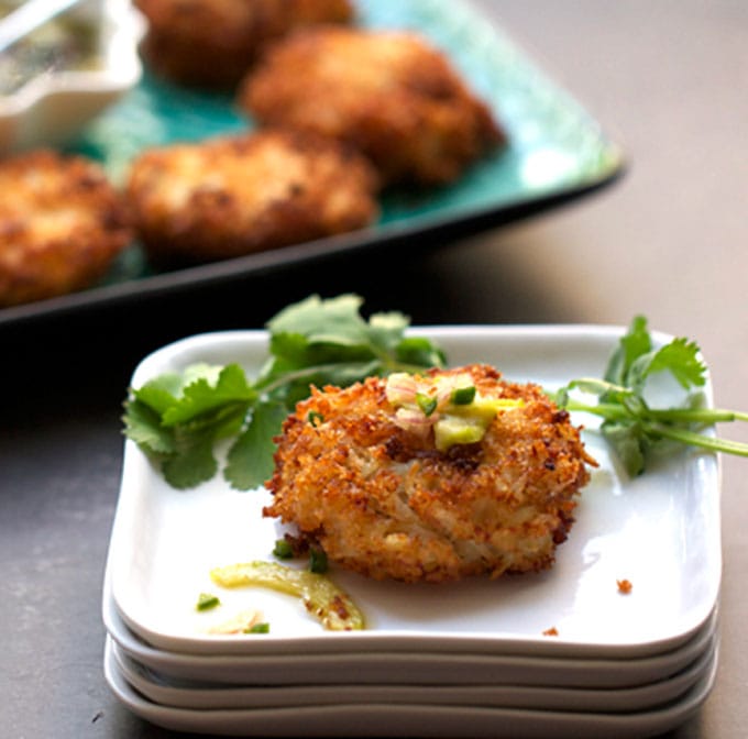 One crab cake on a square appetizer plate with cilantro leave, a platter of crab cakes in the background