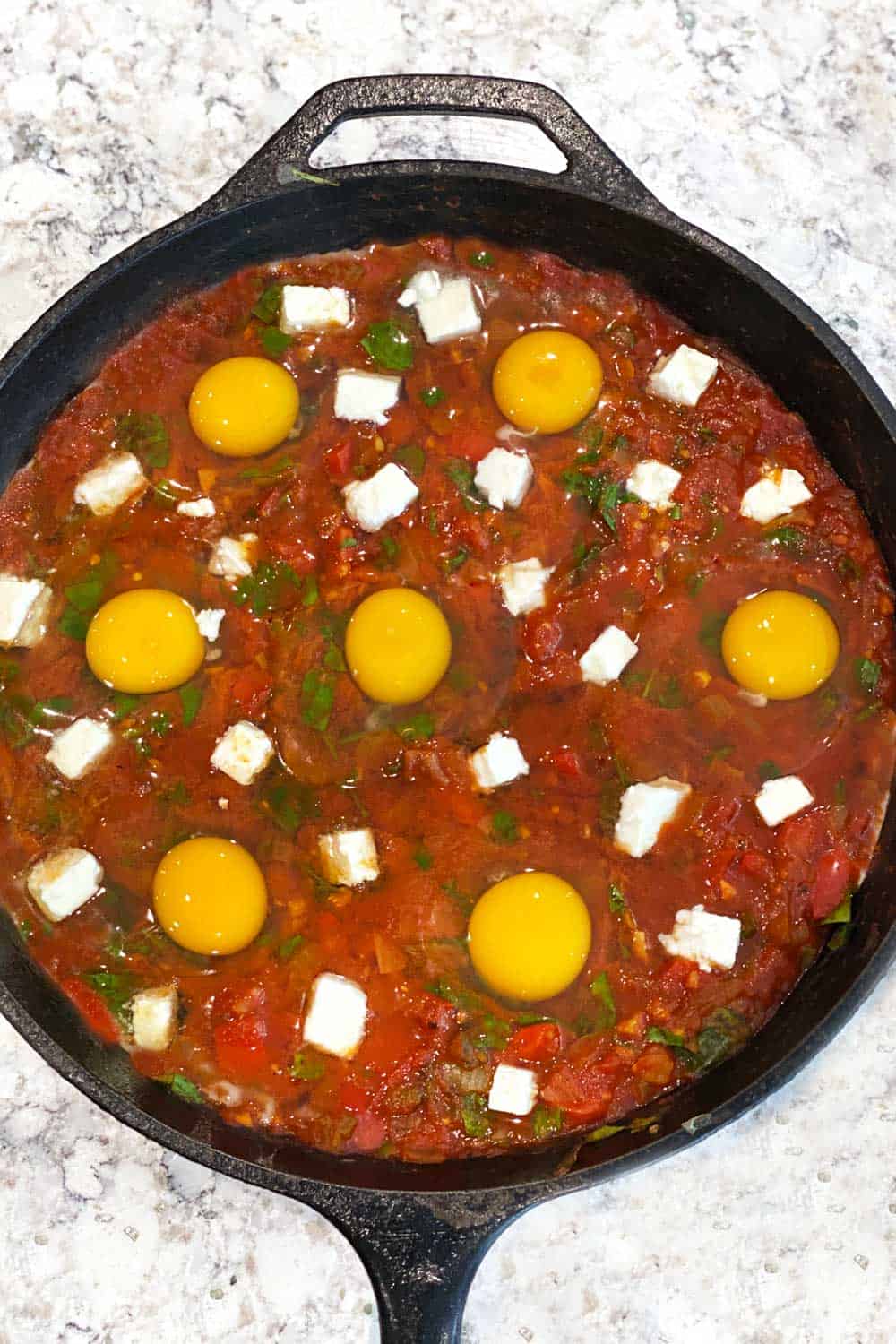 how to make shakshuka, raw eggs and feta in rich thick tomato sauce in a cast iron skillet, ready for the oven