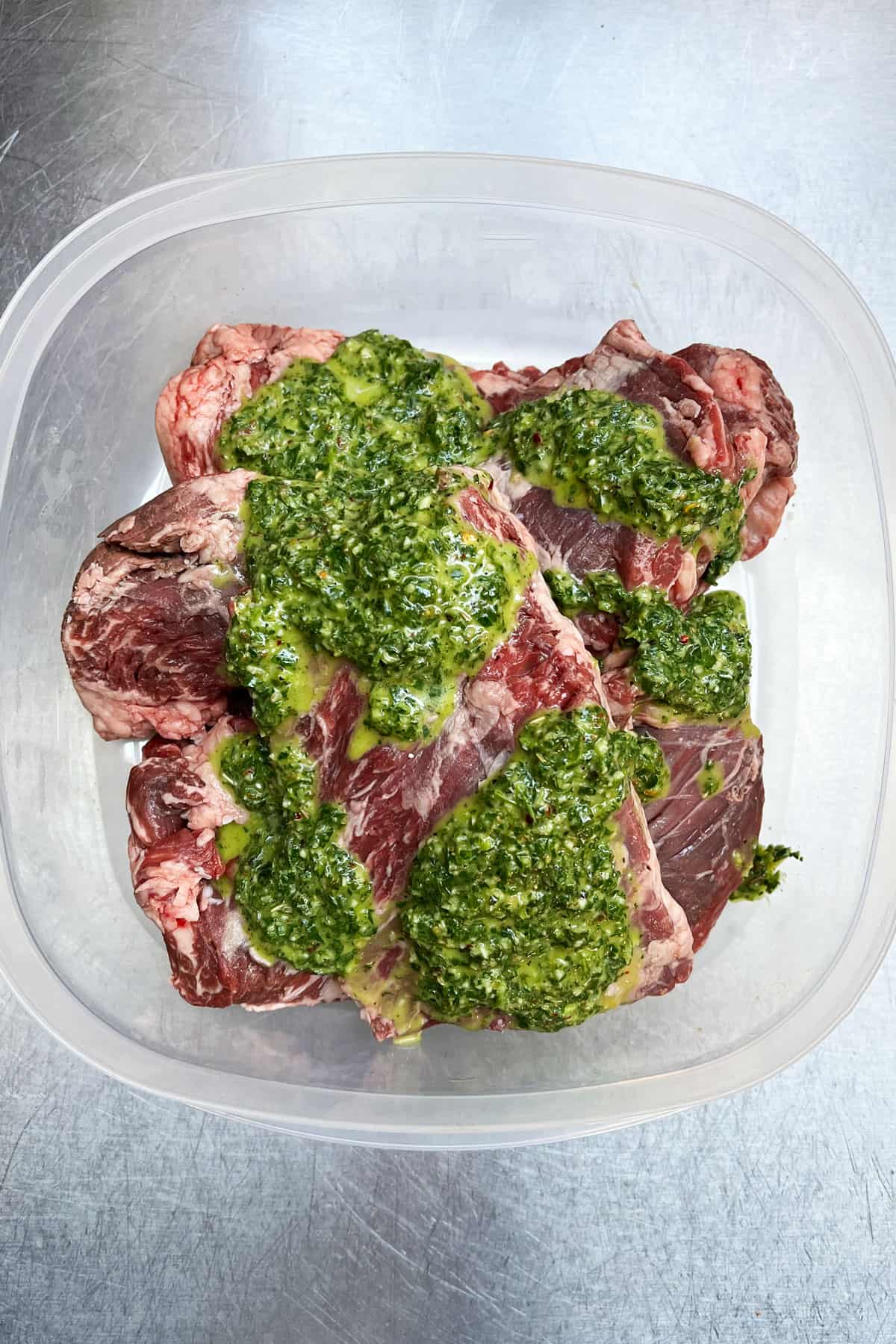 overhead shot of raw skirt steak in a plastic container topped with vibrant green chimichurri sauce marinade