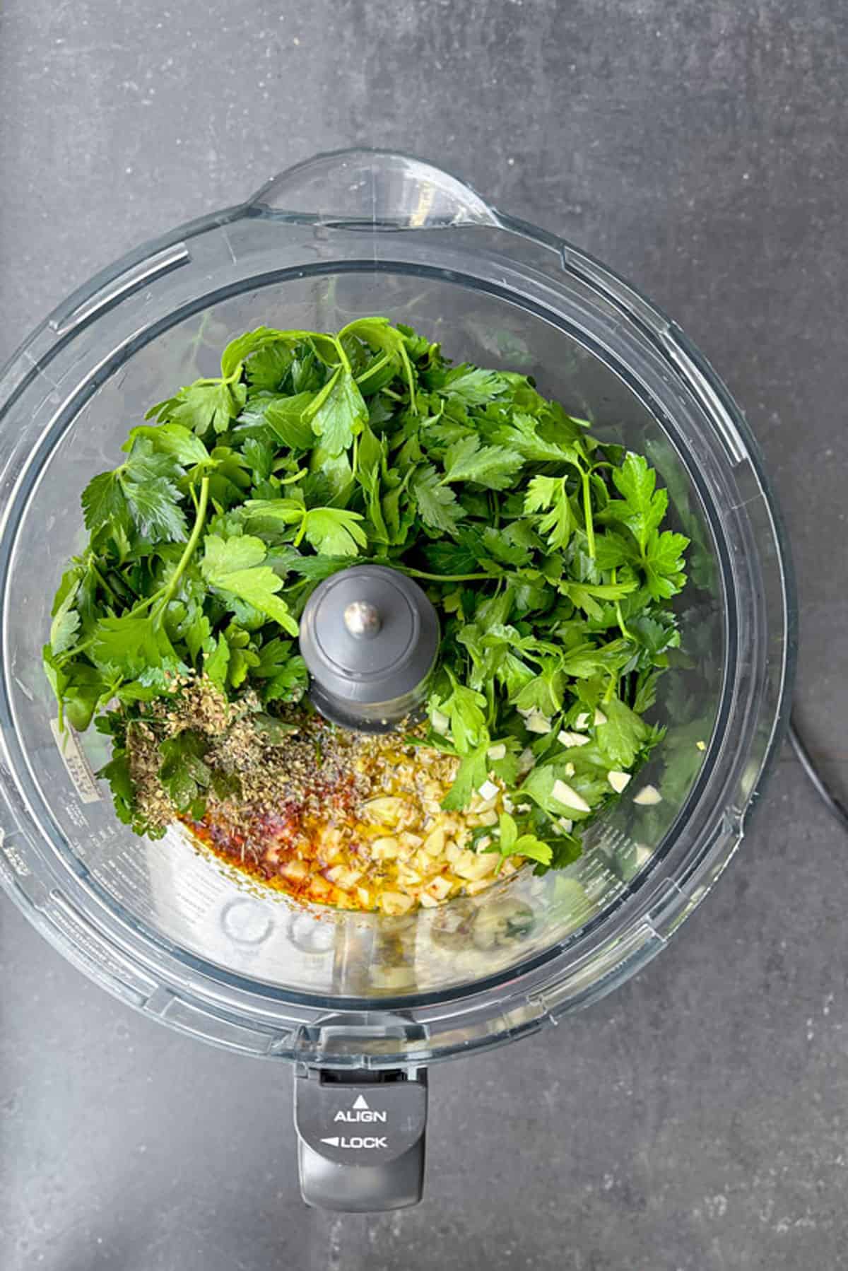 overhead shot of chimichurri ingredients in a food processor: parsley, garlic, oil, vinegar and spices