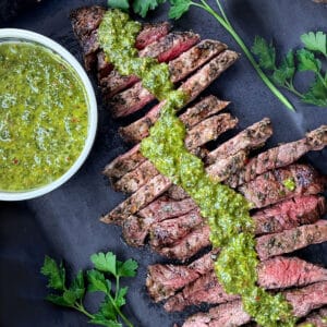 overhead shot of sliced grilled skirt steak arranged diagonally across a dark grey serving board with a line of vivid green chimichurri sauce down the middle and a bowl of chimichurri on the left