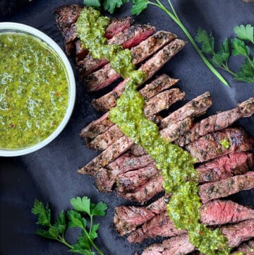overhead shot of sliced grilled skirt steak arranged diagonally across a dark grey serving board with a line of vivid green chimichurri sauce down the middle and a bowl of chimichurri on the left