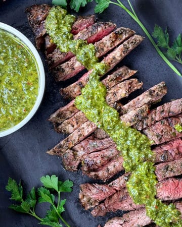 overhead shot of sliced grilled skirt steak arranged diagonally across a dark grey serving board with a line of vivid green chimichurri sauce down the middle and a bowl of chimichurri on the left
