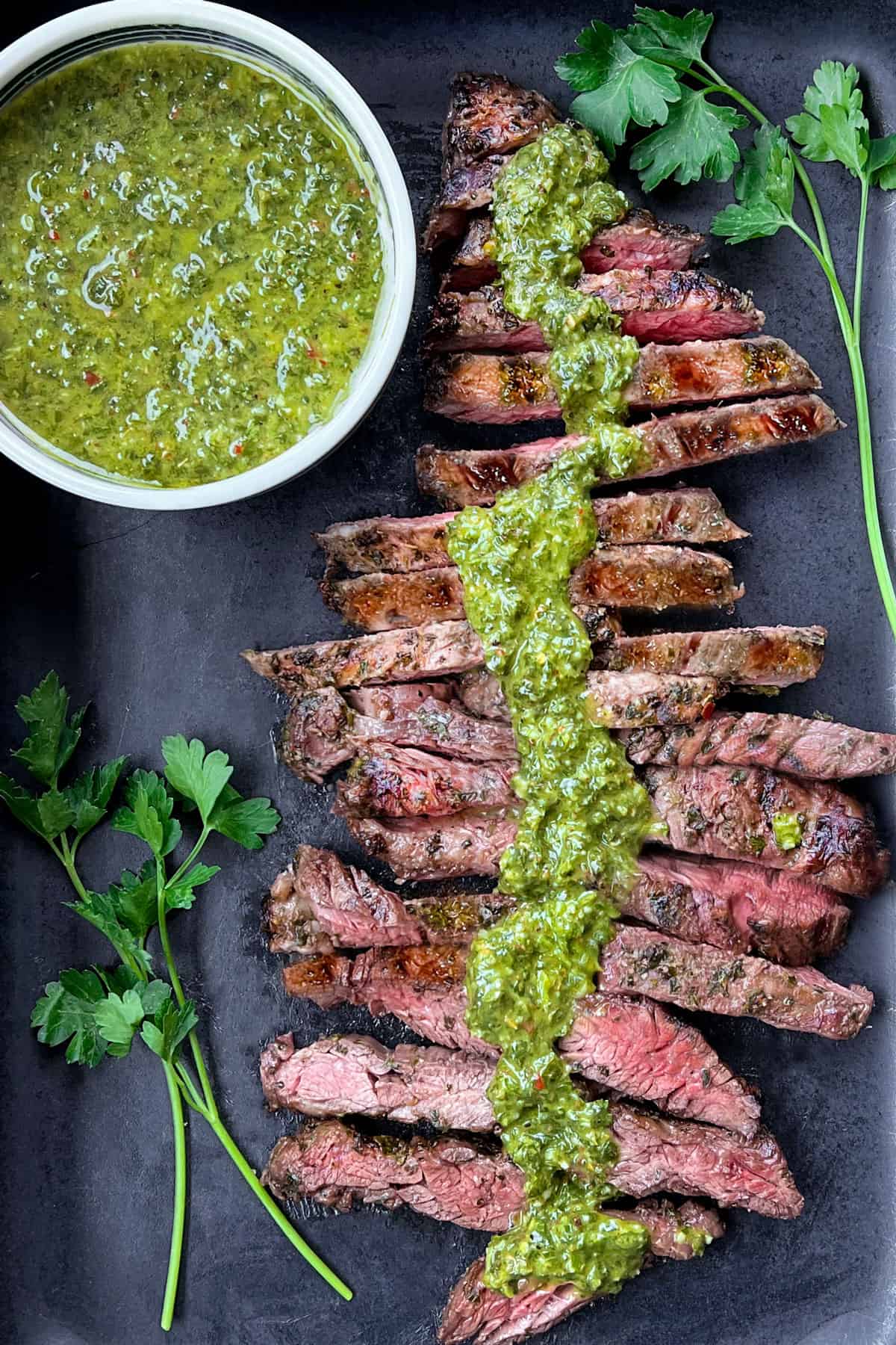overhead shot of a sliced skirt steak, medium rare, with a vertical strip of chimichurri sauce down the middle and a small bowl of the sauce at the upper left