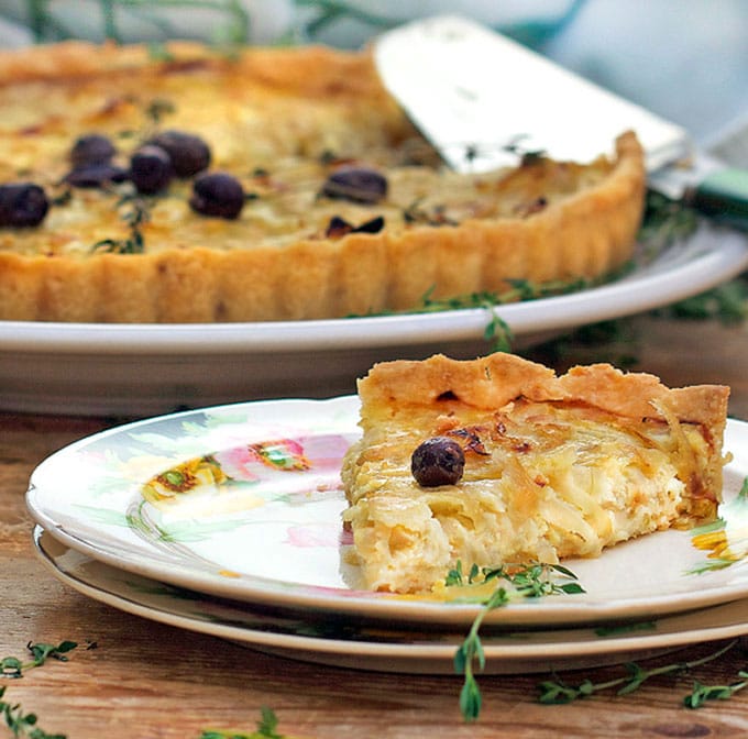 a china plate topped with a triangular slice of a French onion tart with a nicoise olive on top, the rest of the tart in the background.