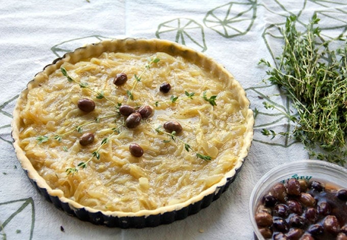 An unbaked French onion tart filled with braised onions and topped with nicoise olives and 6 thyme sprigs, ready to go in the oven