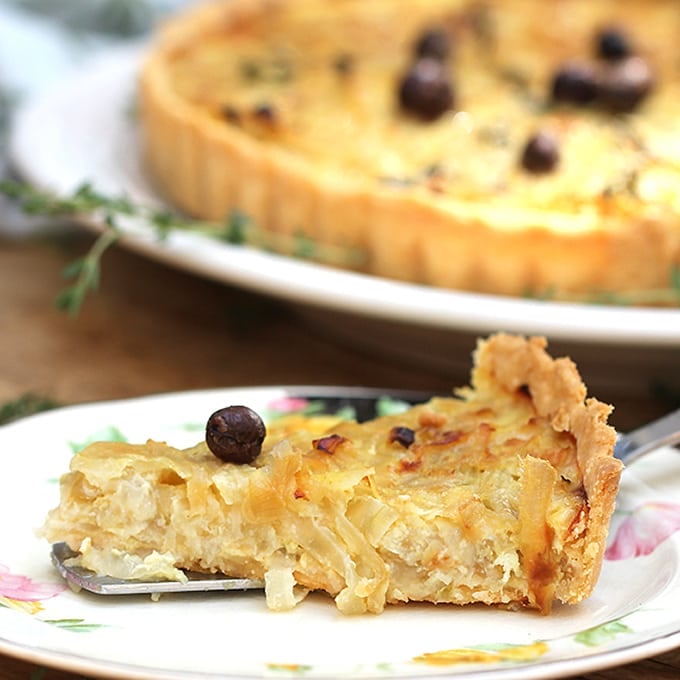 Close up of a slice of French onion tart with a nicoise olive on top, the rest of the tart in the background