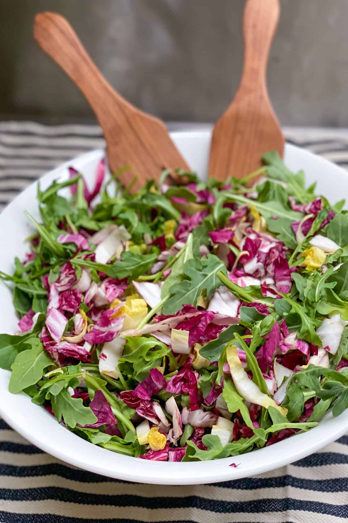 White bowl on a black and white striped dish towel, filled with tricolore salad: arugula, radicchio and endive, with wooden salad spoons in the bowl.