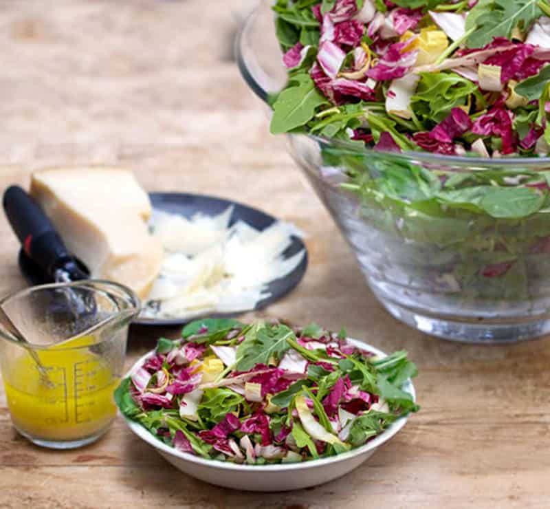 Tricolore salad in a small bowl with a measuring cup filled with lemon vinaigrette and a small black plate topped with a hunk of parmigiana and some parmesan shavings 