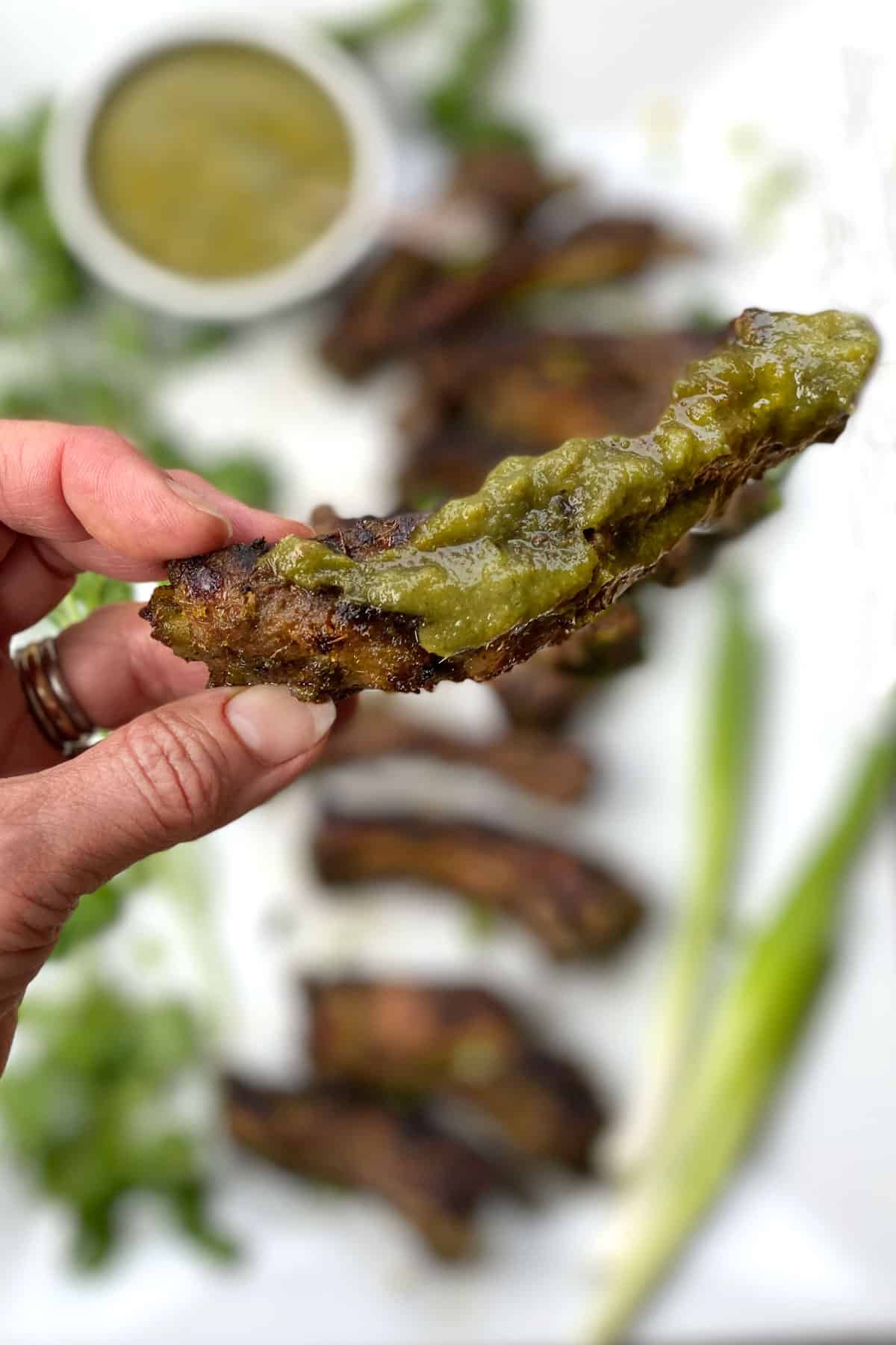 fingers holding one Thai BBQ rib coated with green bbq sauce, a platter of ribs in the background