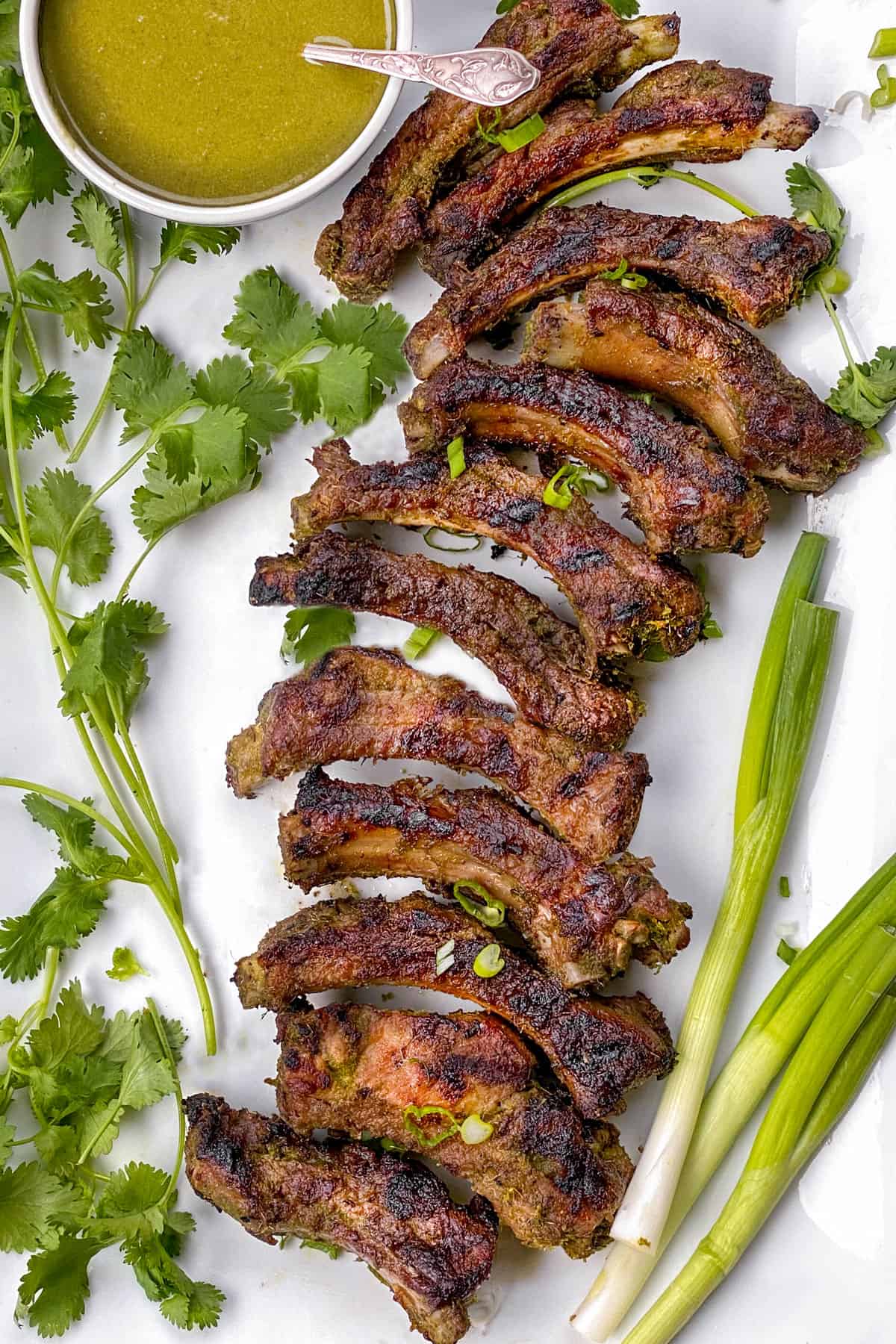 A rack of separated grilled baby back ribs on a white platter with sprigs of cilantro on the left, a few green onions on the right, and a bowl of green Thai bbq sauce at the top left